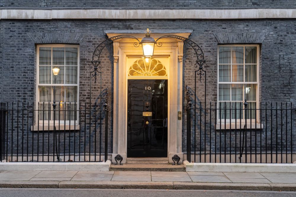 Photo shows the front door of number ten downing street - the door is oak and painted black, it is flanked by ornate cast iron railings and an arch between which a victorian style lamp is lit.