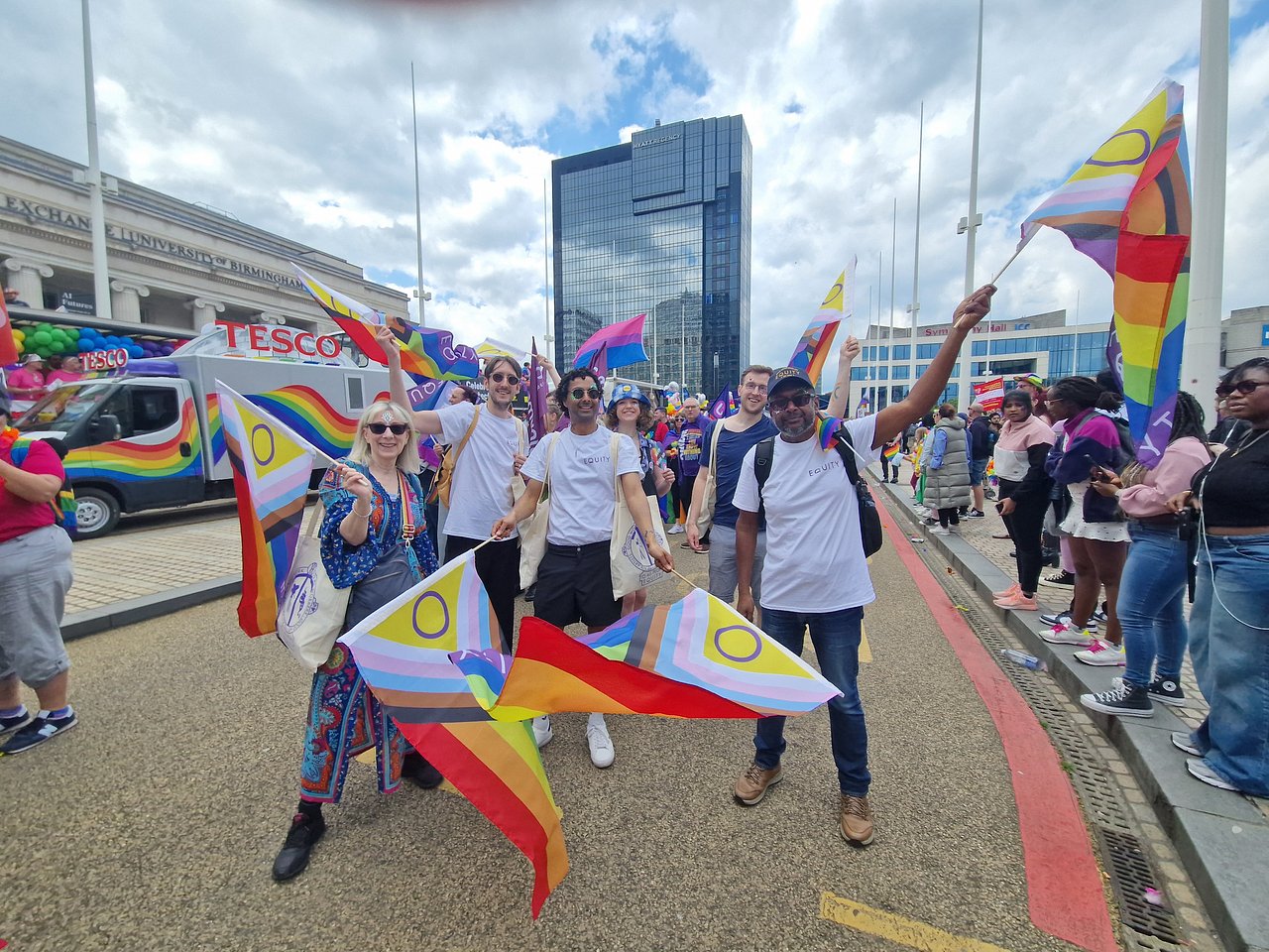 A group of six Equity members in Birmingham city centre. They are holding pride flags and all smiling at the camera. The parade is going on around them, and there are people standing on the pavement to one side watching.