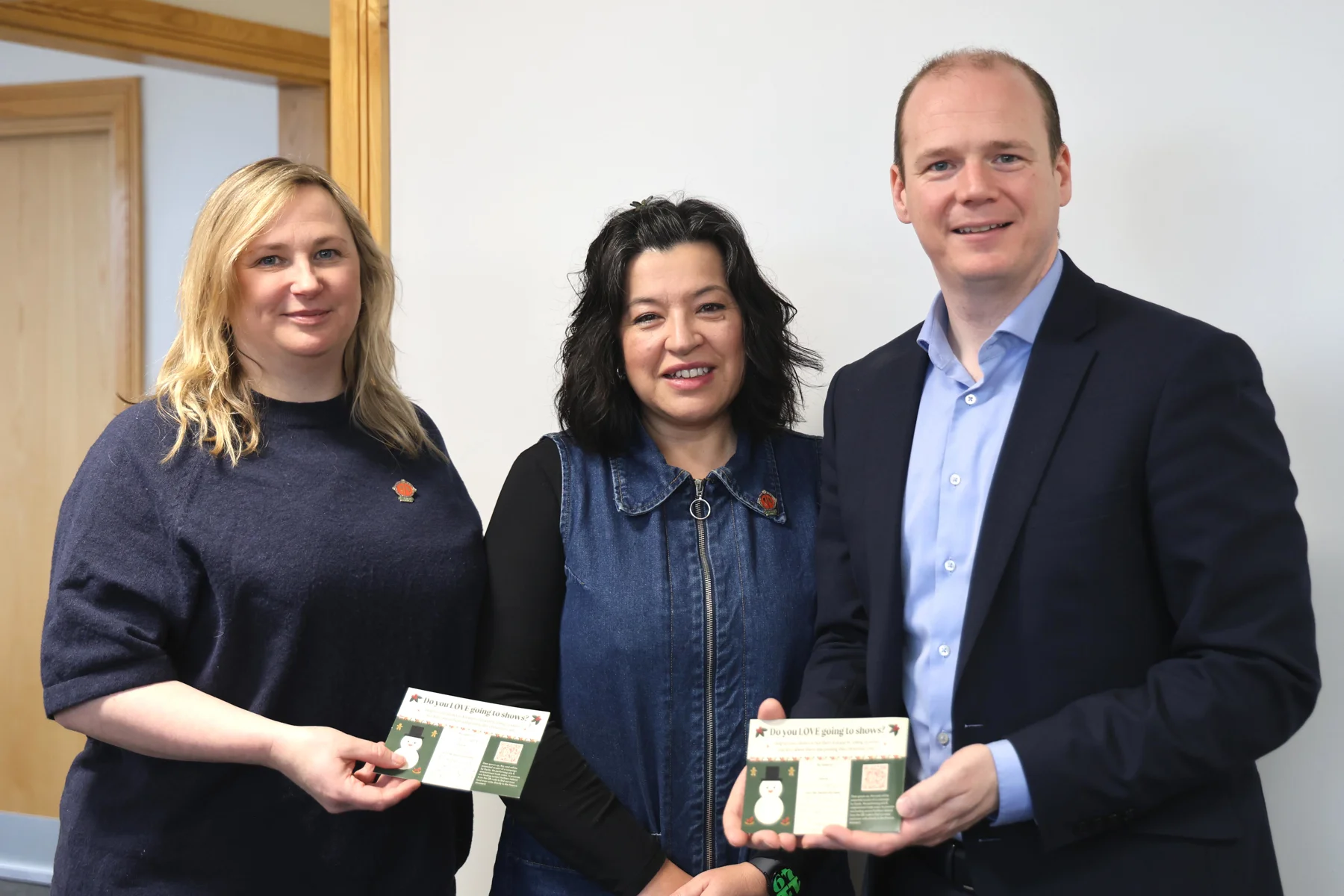 Two women and a man hold two Christmas postcards that read 'Do you LOVE going to shows'.