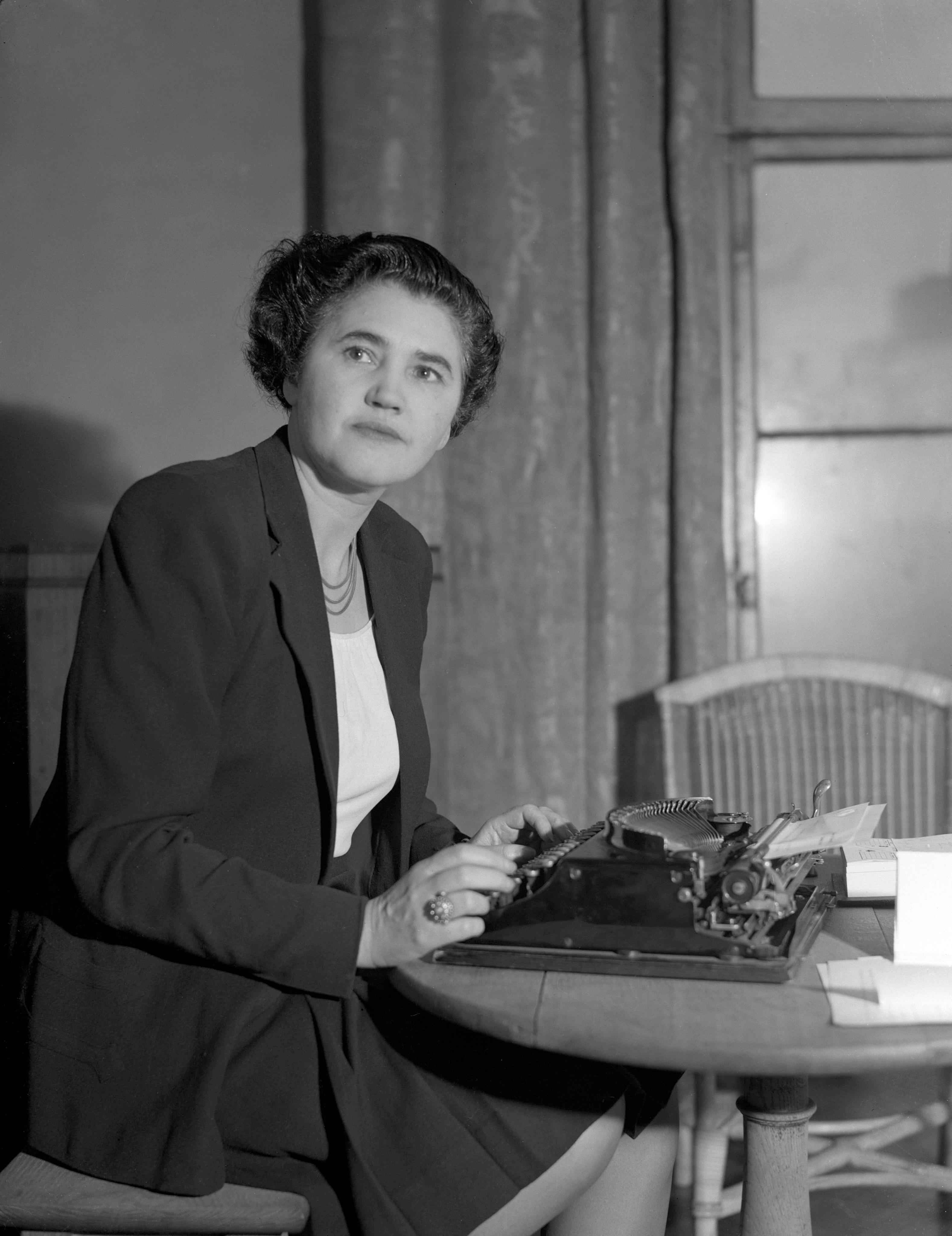 Jennie Lee is sat at a desk writing on a type writer, a black and white photo. 